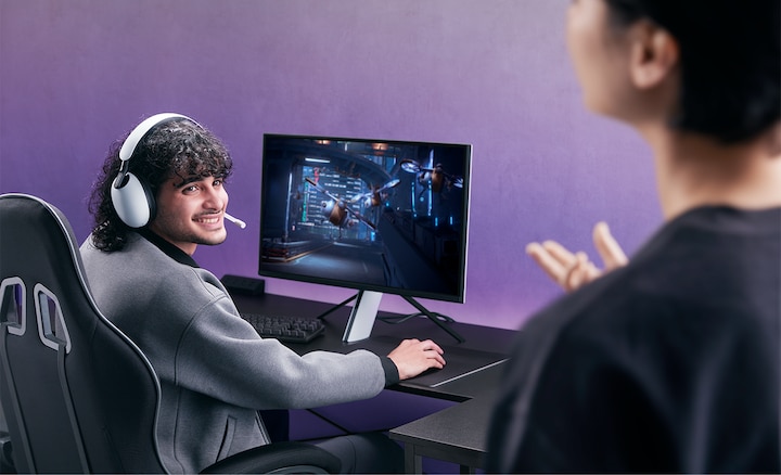 Man wearing INZONE H9 headset sitting at a desk with monitor and game on screen, talking to partner in foreground right