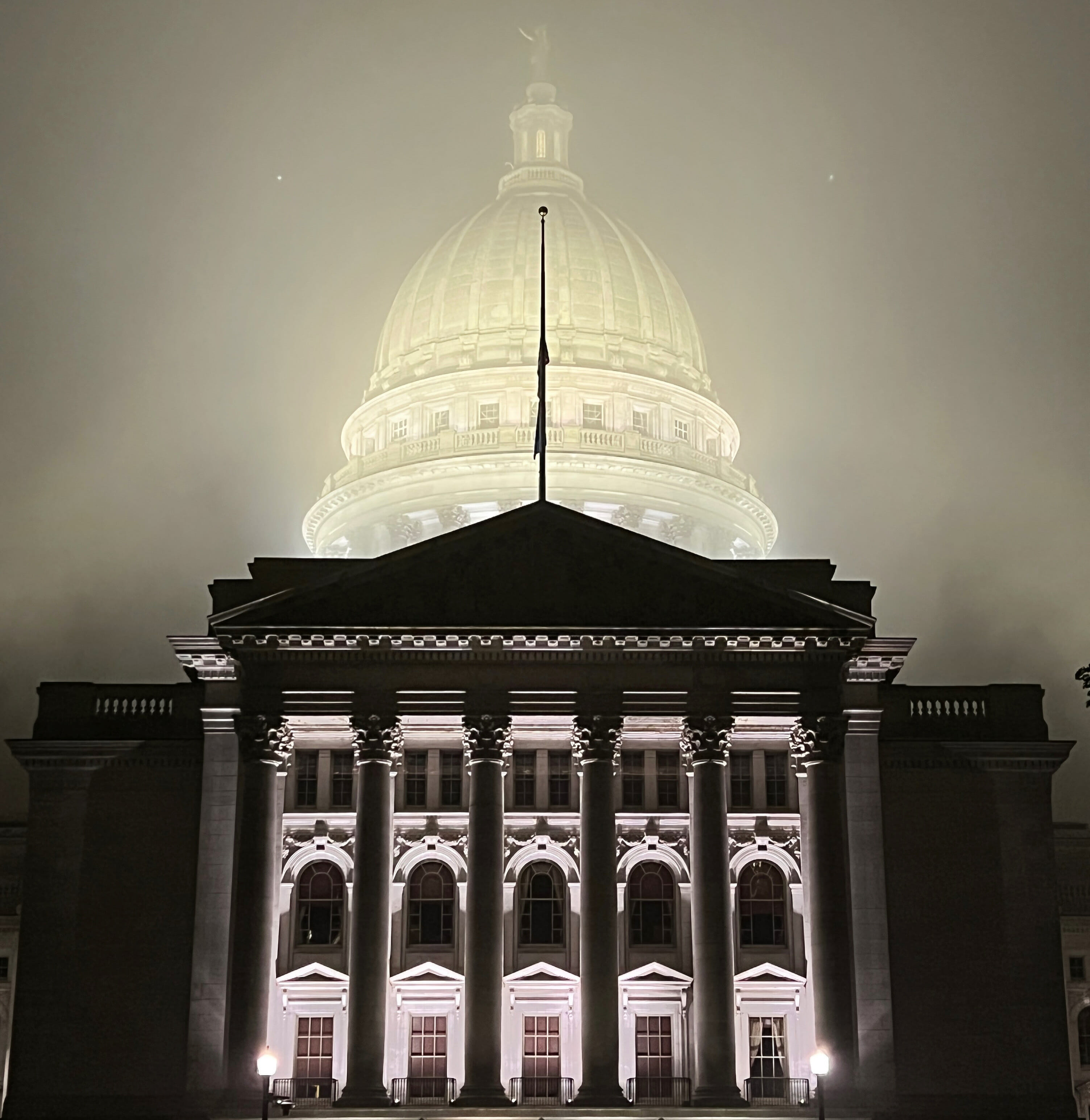 Stark columns of the Wisconsin State Capitol building are clear in the foreground while the lit-up dome behind is blurred by fog.