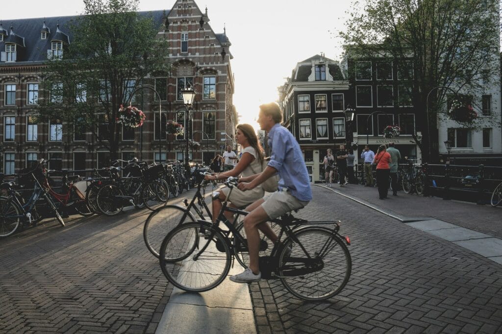 White man in a blue shirt and white woman with braided hair wearing a white dress riding bicycles through a city-setting