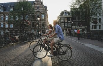 White man in a blue shirt and white woman with braided hair wearing a white dress riding bicycles through a city-setting